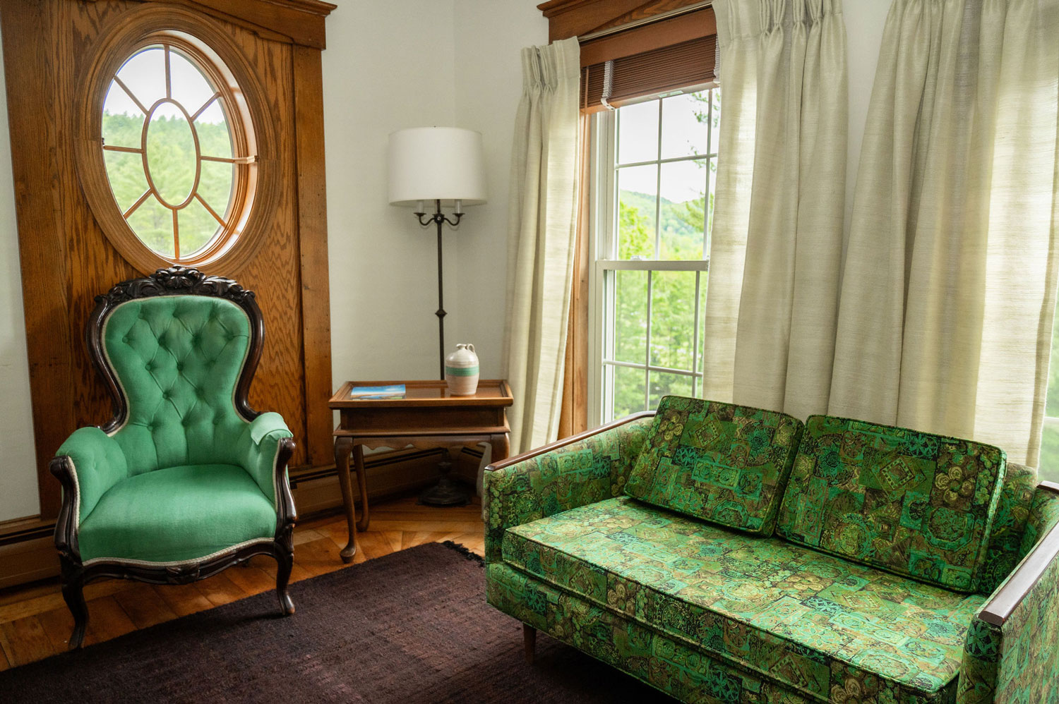 A green upholstered chair underneath an oval window across from a matching love seat.