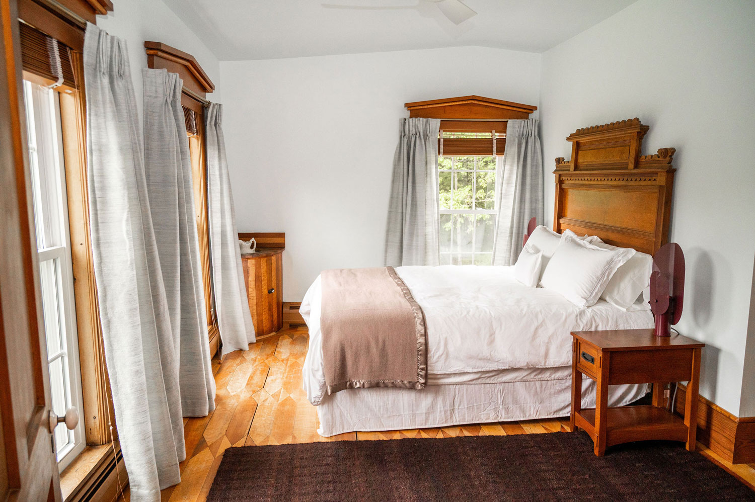 The Sparhawk Bedroom with a bed with carved headboard, a corner cabinet with an old fashioned pitcher and basin, and large window.