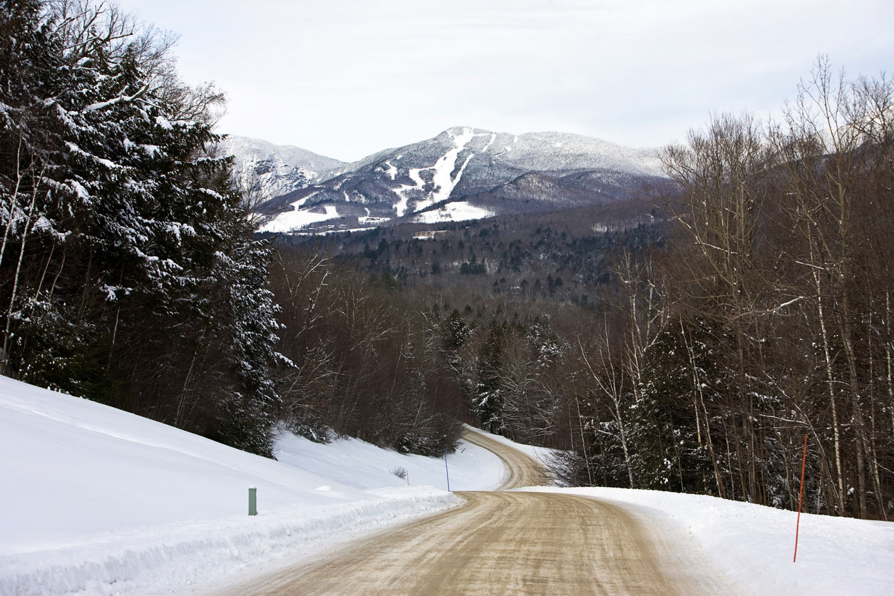 A winter road passing through snowy forest with a ski slopes visible on a hill in the distance.